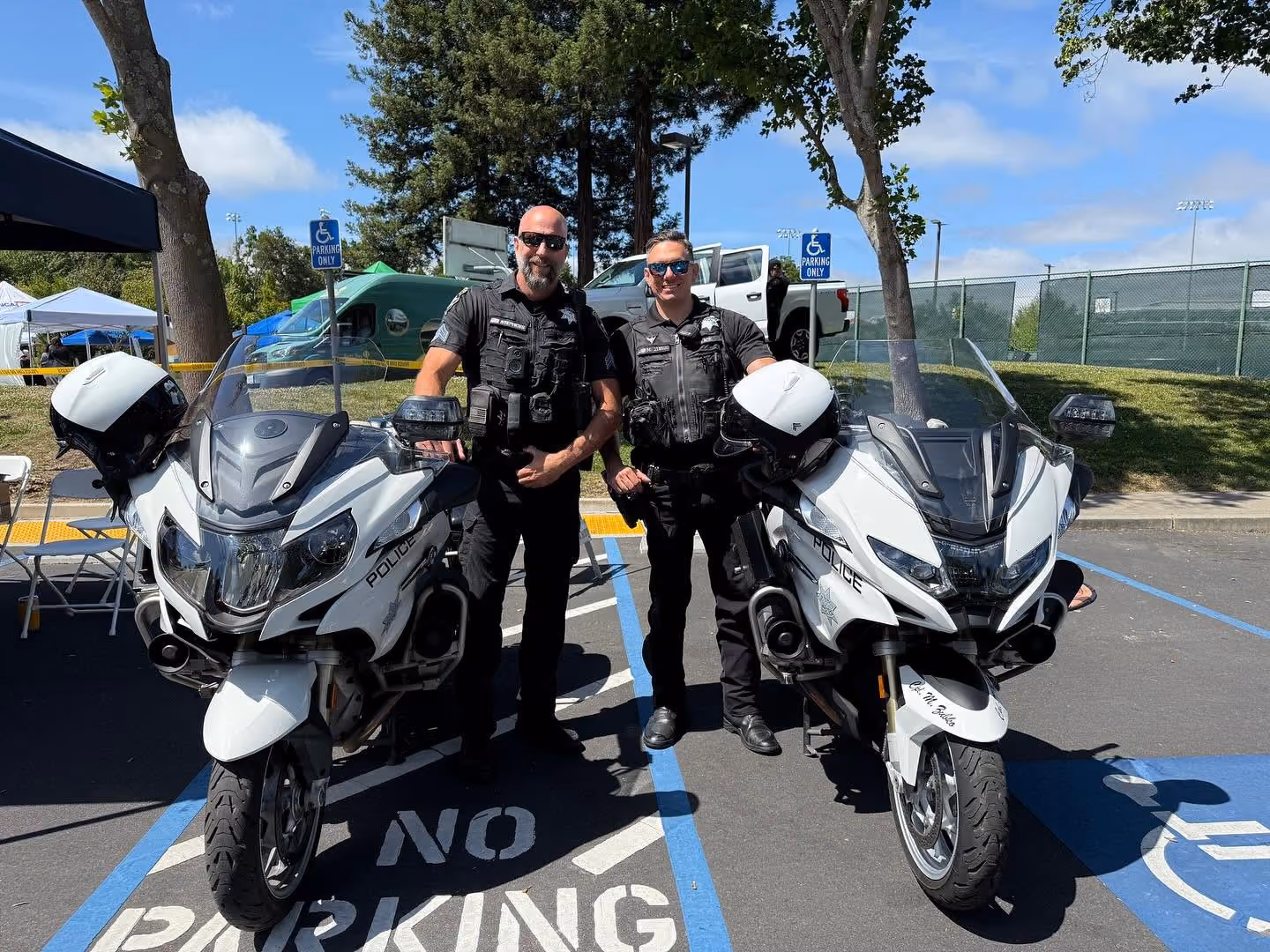 Two police officers in uniform standing next to their white police motorcycles in a parking lot on a sunny day with trees and tents in the background.