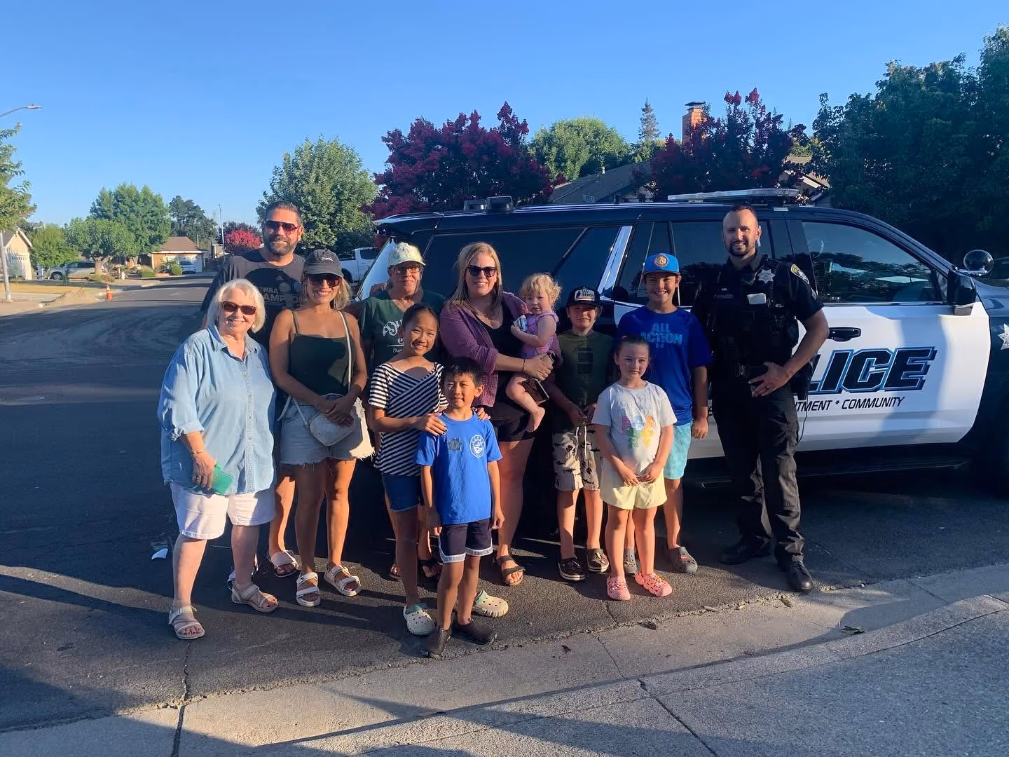 Group of adults, children, and a police officer standing together on a sunny street in front of a black and white police SUV with visible markings, smiling at the camera.
