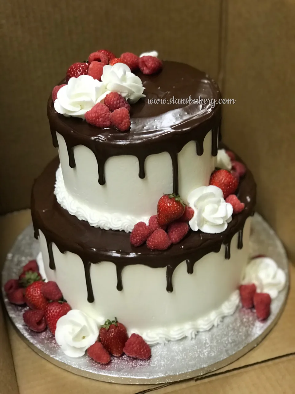 A two-tier white cake with a chocolate drip, fresh raspberries, strawberries and piped roses
