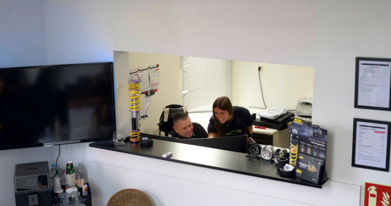 Empfangsbereich mit Theke und großen Fensteröffnung, durch die zwei Personen an einem Computer lachen und zusammenarbeiten; auf der Theke Technikzubehör und Produktinformationen sichtbar.