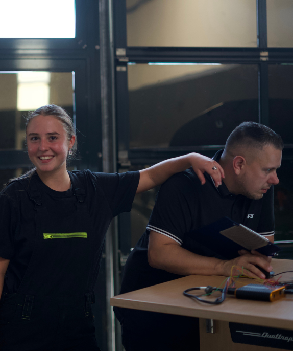 Junge Frau lächelt und legt ihre Hand freundschaftlich auf die Schulter eines Mannes, der an einem Tisch mit elektronischen Geräten und einem Notizblock arbeitet.