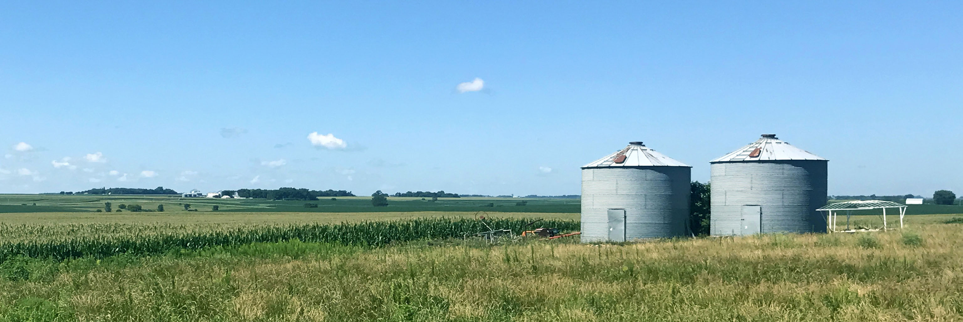 Two metal grain silos stand in a grassy field under a blue sky with distant farmland.