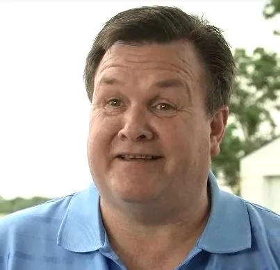 Smiling middle-aged man with brown hair wearing a light blue collared shirt outdoors.