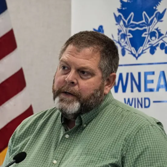 Man with beard wearing green checked shirt speaking in front of flag and blue logo on wall.
