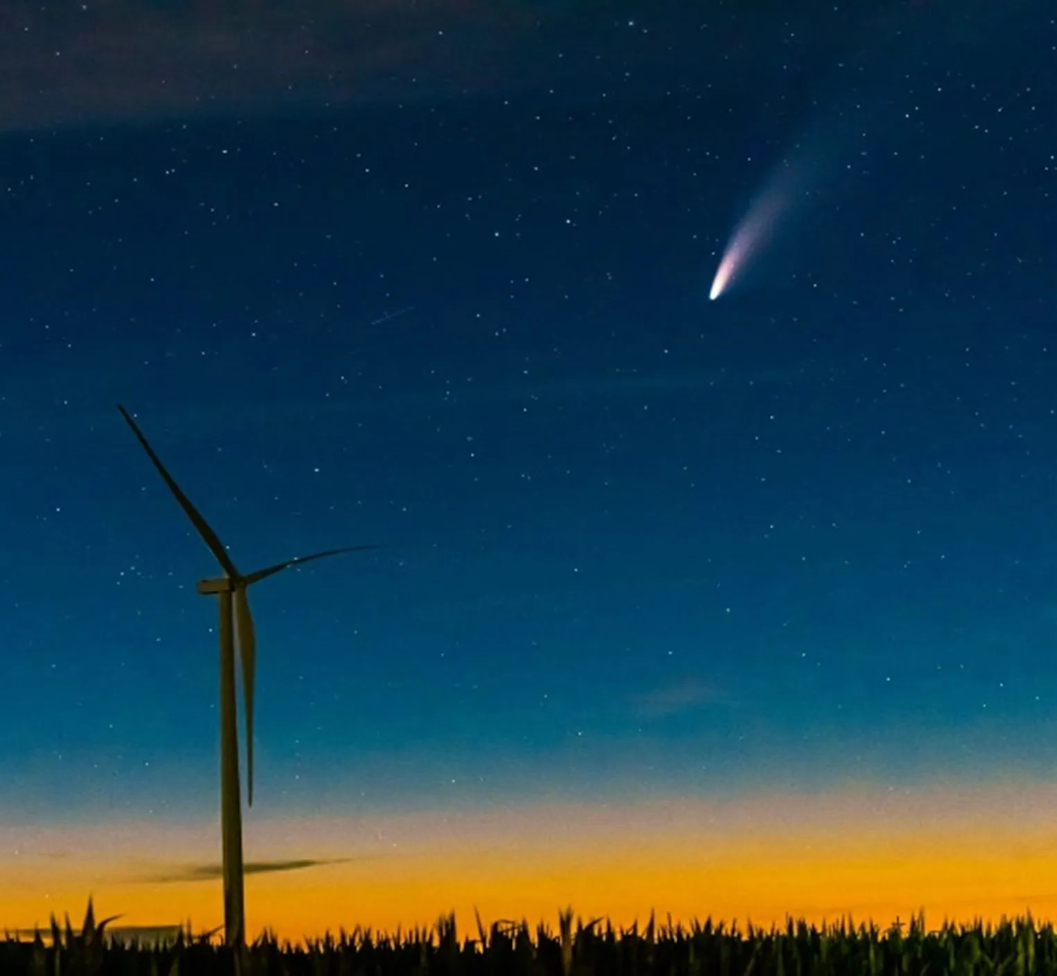 A wind turbine silhouette stands against a starry night sky with a comet streaking above at dusk, with an orange horizon and dark foreground vegetation.