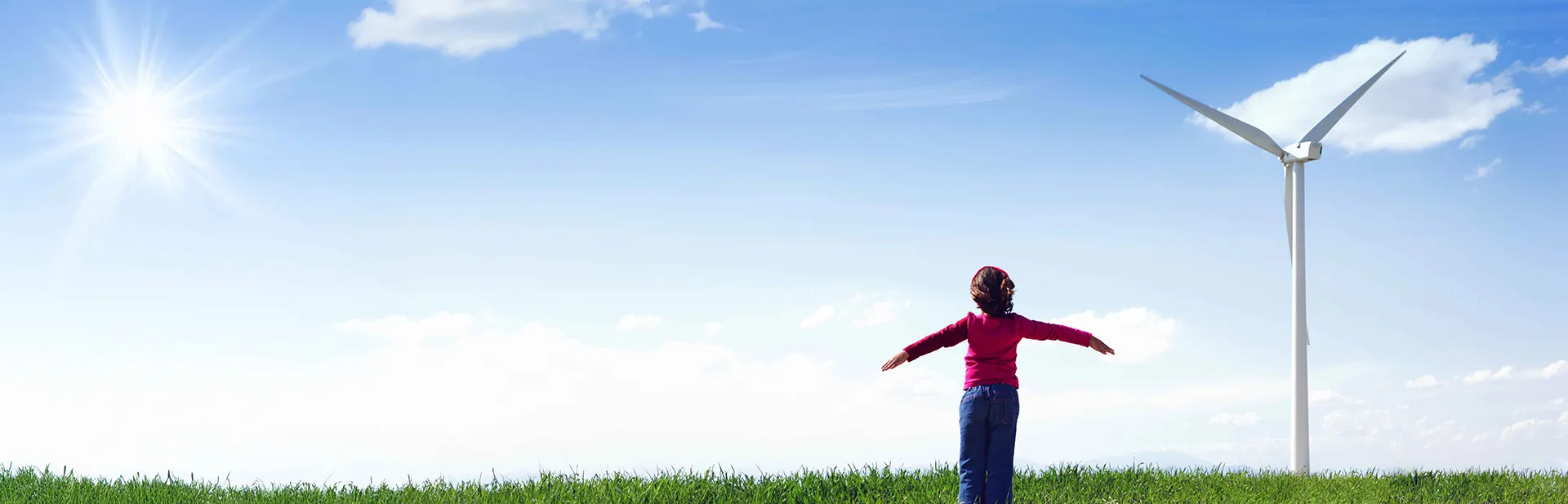 Child with arms outstretched standing on green grass under a bright blue sky with the sun shining, near a large wind turbine.