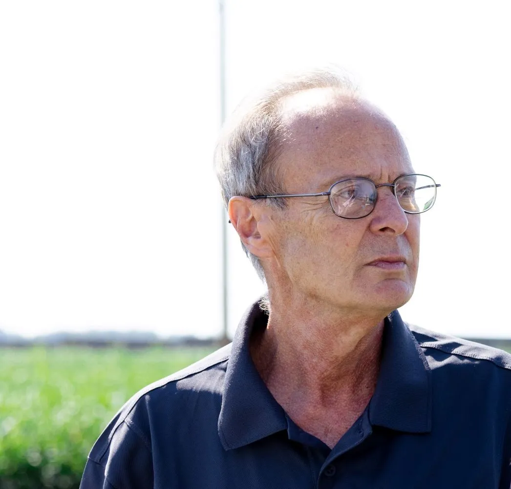 Older man wearing glasses and a dark polo shirt, looking to the right with a green field background.