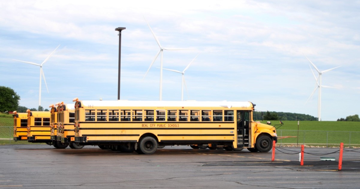 Yellow school buses parked in a lot with green fields and several white wind turbines in the background under a partly cloudy sky.