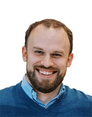 Smiling man with short brown hair, a beard, and blue eyes, wearing a blue sweater over a blue checkered shirt, against a white background.