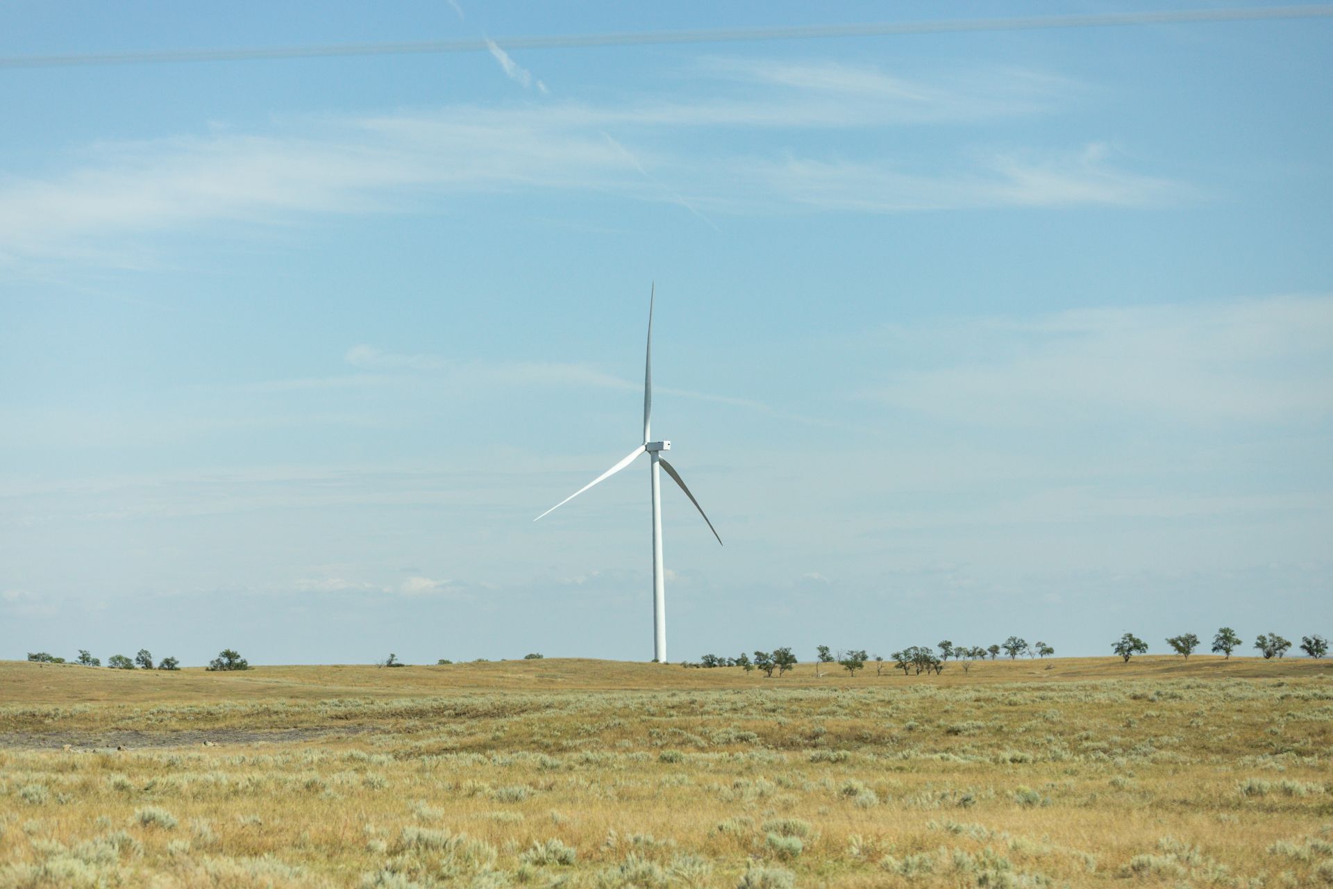 A single white wind turbine stands on a dry grassy plain with sparse small trees under a blue sky with wispy clouds.