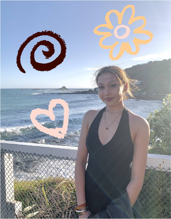 Young woman with pulled-back hair and layered necklaces wearing a black halter dress, standing by a metal fence with ocean and cliffs in the background under a clear sky, with orange doodles of a flower, a heart, and a spiral above her.