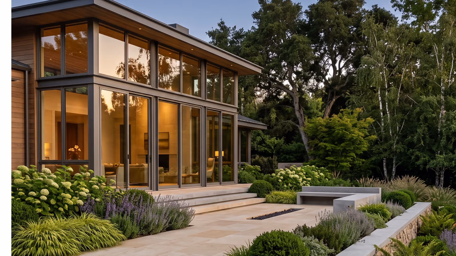 Modern house exterior with large floor-to-ceiling windows reflecting trees, surrounded by lush greenery, manicured shrubs, and a stone patio with built-in concrete benches.