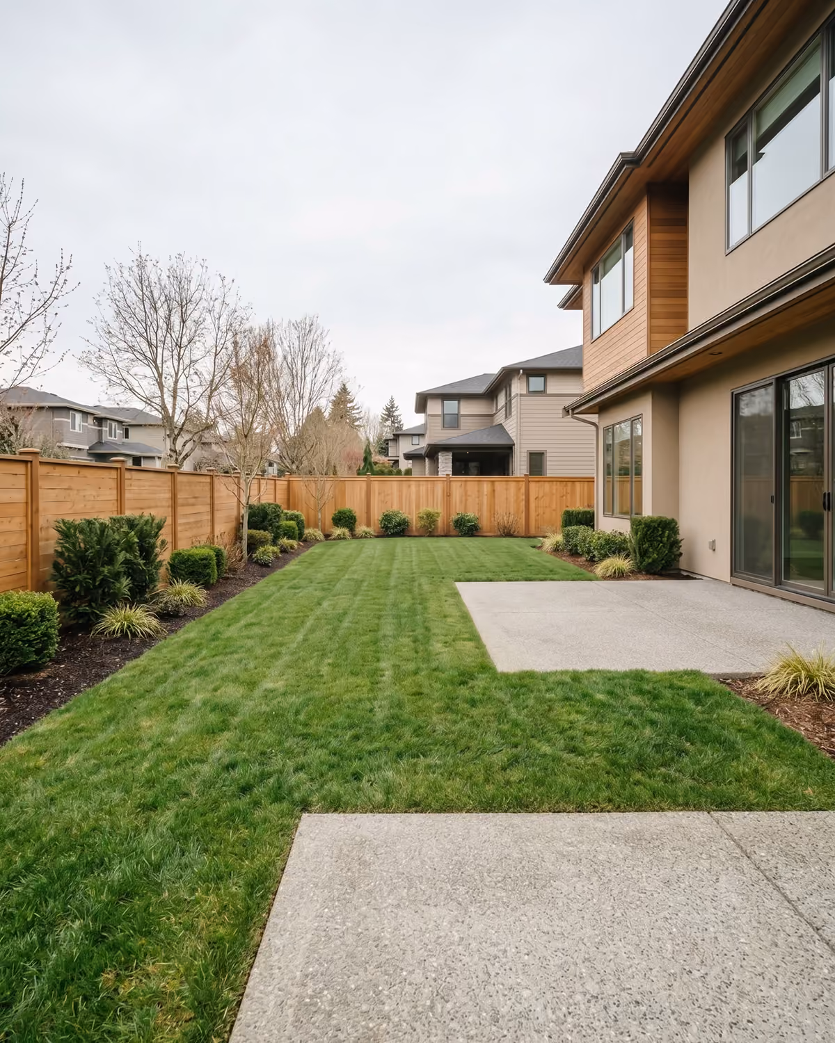 Manicured backyard with a striped lawn, concrete patio, and low hedges along a wood fence, backed by a modern home with wood and stucco siding.