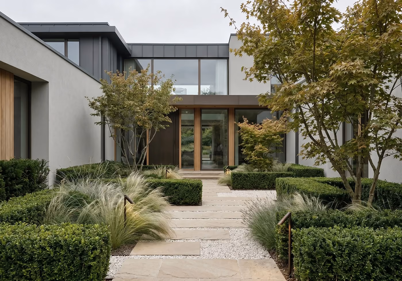 Symmetrical entry courtyard of a modern home with a wood-framed front door, a stone-and-gravel path flanked by clipped hedges, ornamental grasses, and maple trees.