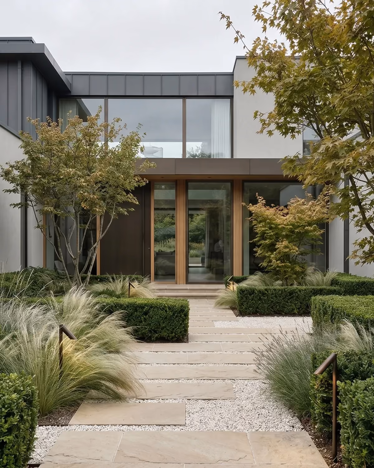 Symmetrical entry courtyard of a modern home with a wood-framed front door, a stone-and-gravel path flanked by clipped hedges, ornamental grasses, and maple trees.