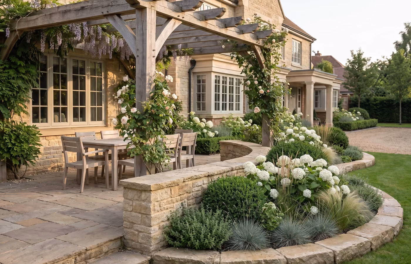 Stone patio under a wooden pergola draped in wisteria and climbing roses, with a wood dining set and a curved retaining wall bordering a bed of white hydrangeas and ornamental grasses, set against a natural stone country house.