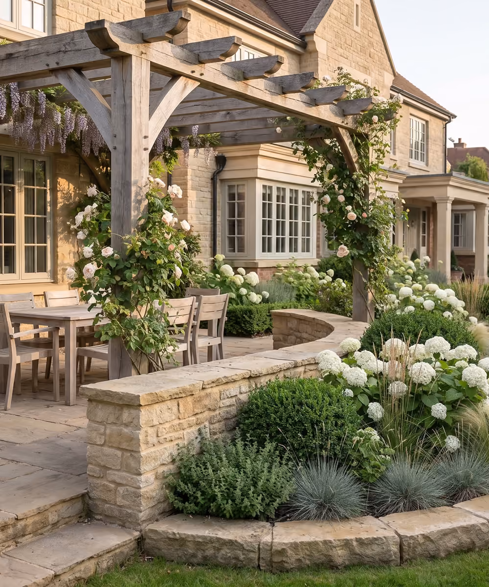 Stone patio under a wooden pergola draped in wisteria and climbing roses, with a wood dining set and a curved retaining wall bordering a bed of white hydrangeas and ornamental grasses, set against a natural stone country house.