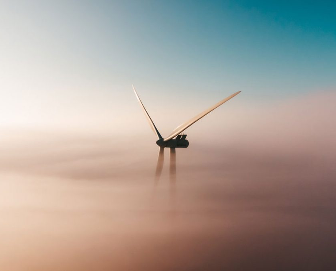 Two wind turbines partially submerged in a thick layer of mist or fog, with a clear blue sky above.
