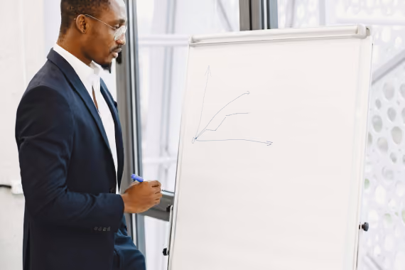 Man in suit holding a marker and standing next to a flip chart with a simple line graph drawn on it.
