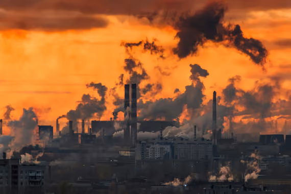 Industrial cityscape at sunset with numerous smokestacks releasing thick black and gray smoke into an orange sky, indicating air pollution.
