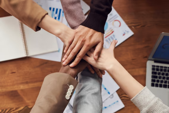 Five people stacking their hands together in a teamwork gesture over a desk with documents and a laptop.