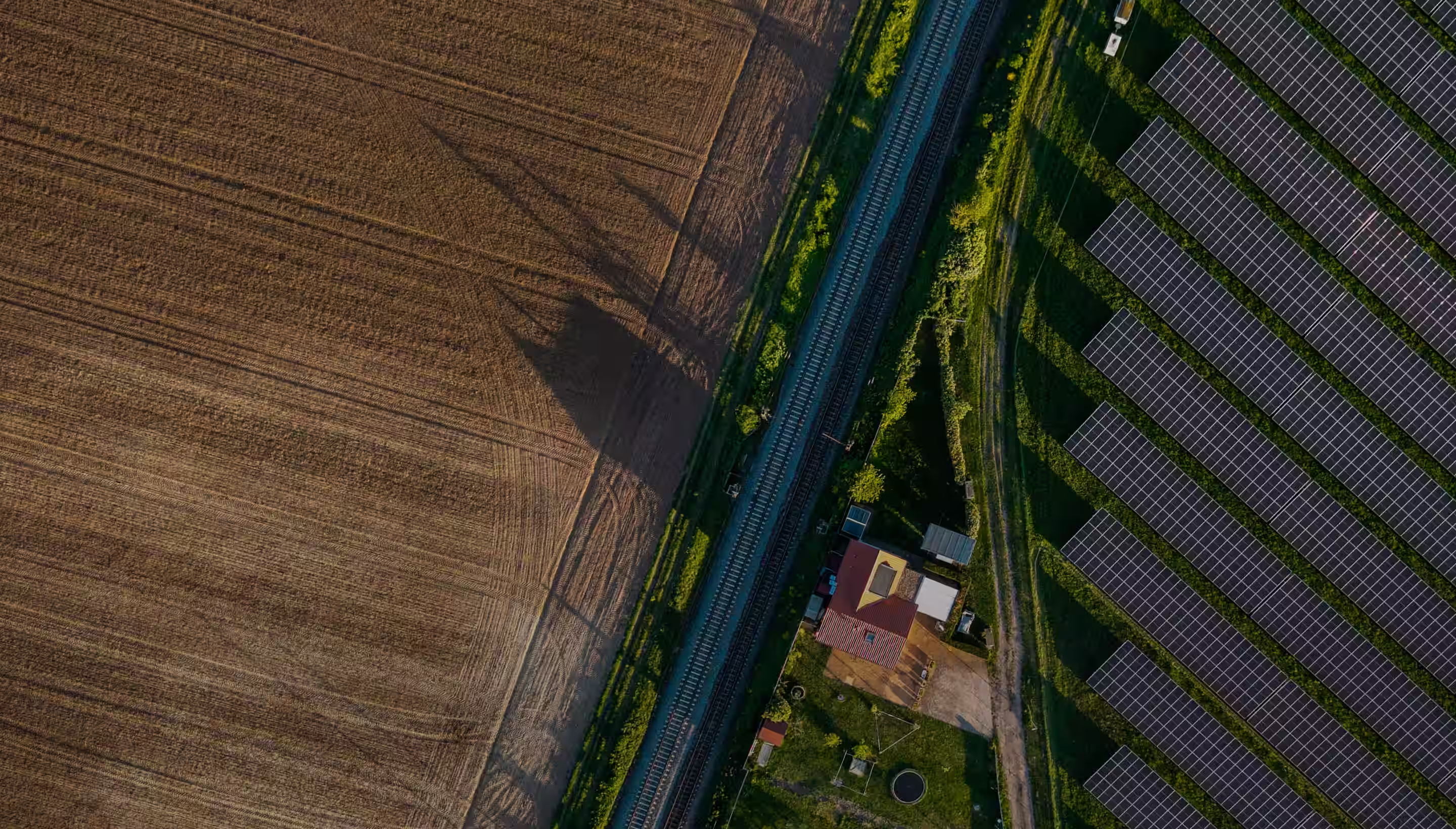 Aerial view showing a field on the left, a railway track running vertically in the center, and rows of solar panels on the right with a small building and greenery below the tracks.