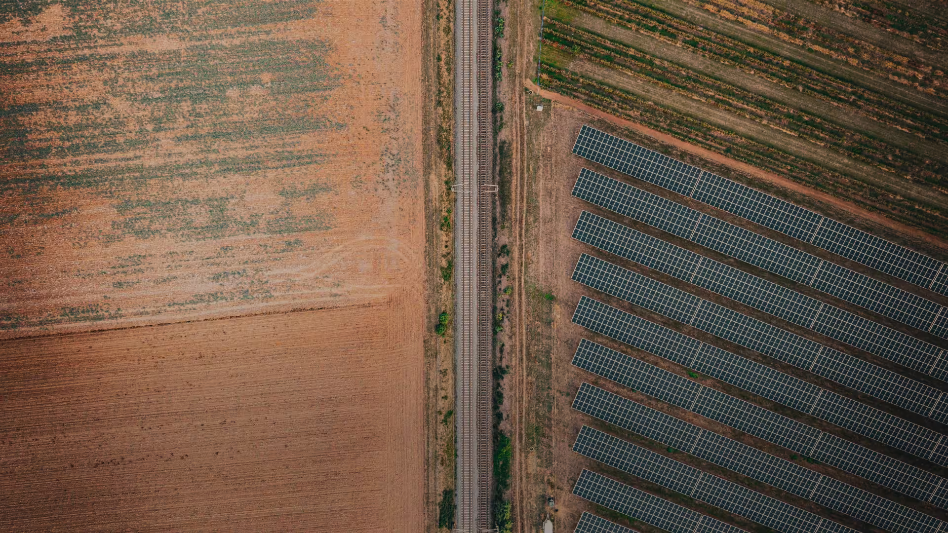 Aerial view showing a field on the left, a railway track running vertically in the center, and rows of solar panels on the right with a small building and greenery below the tracks.
