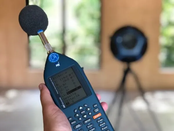 Hand holding a sound level meter with a measurement sound source on a tripod behind