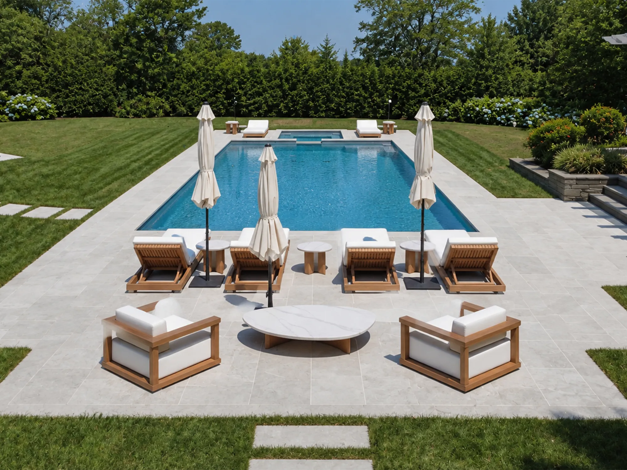 Light stone paving around a garden swimming pool, with wooden loungers, white umbrellas, and an outdoor seating area.