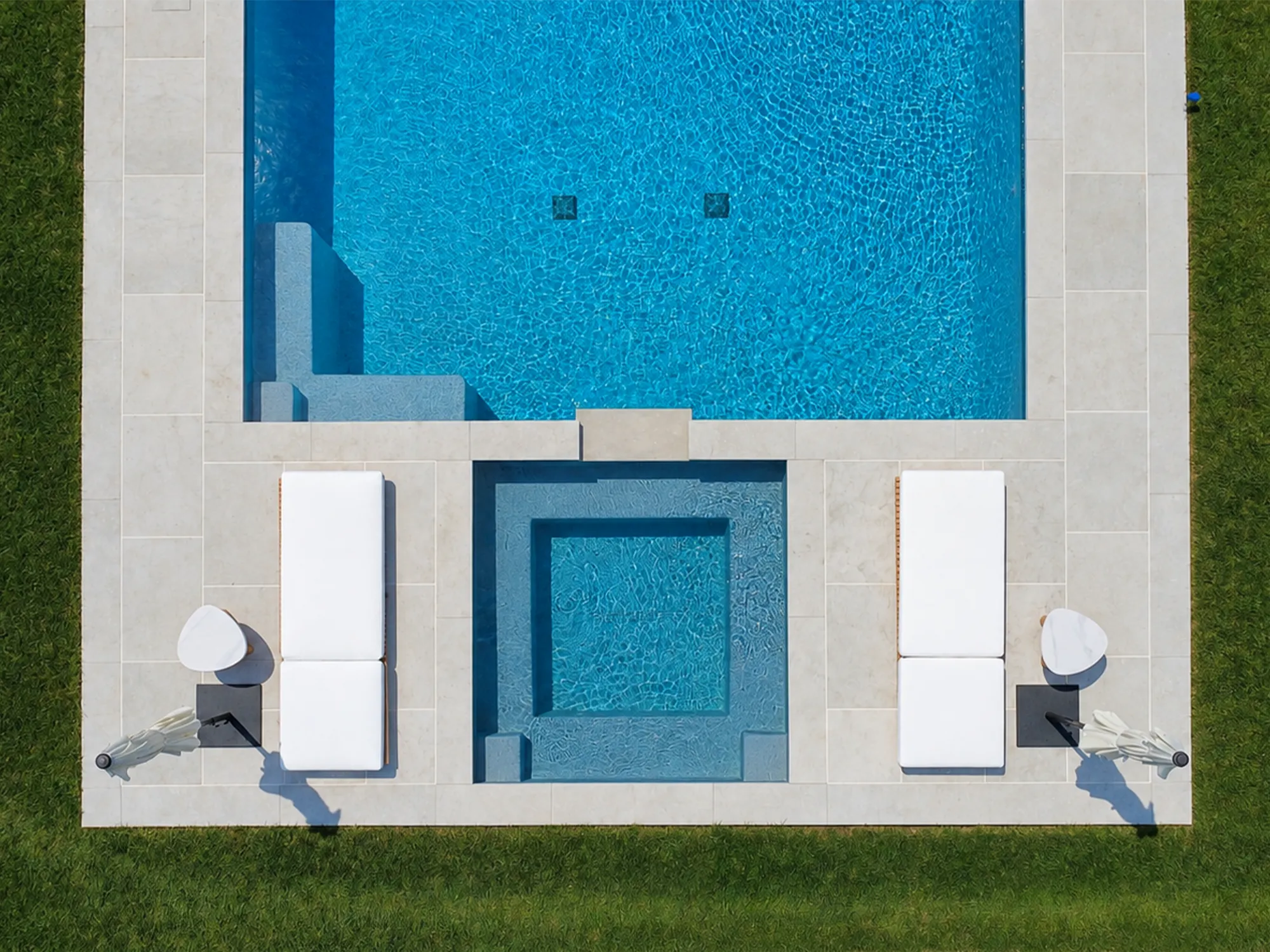 Overhead view of light stone paving around a swimming pool and square spa, with two white loungers.
