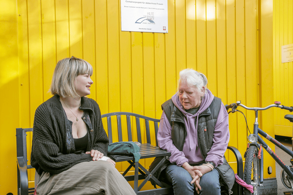 Zwei Frauen im Gespräch auf einer Bank vor einer gelben Wand im Containerprojekt der Caritas
