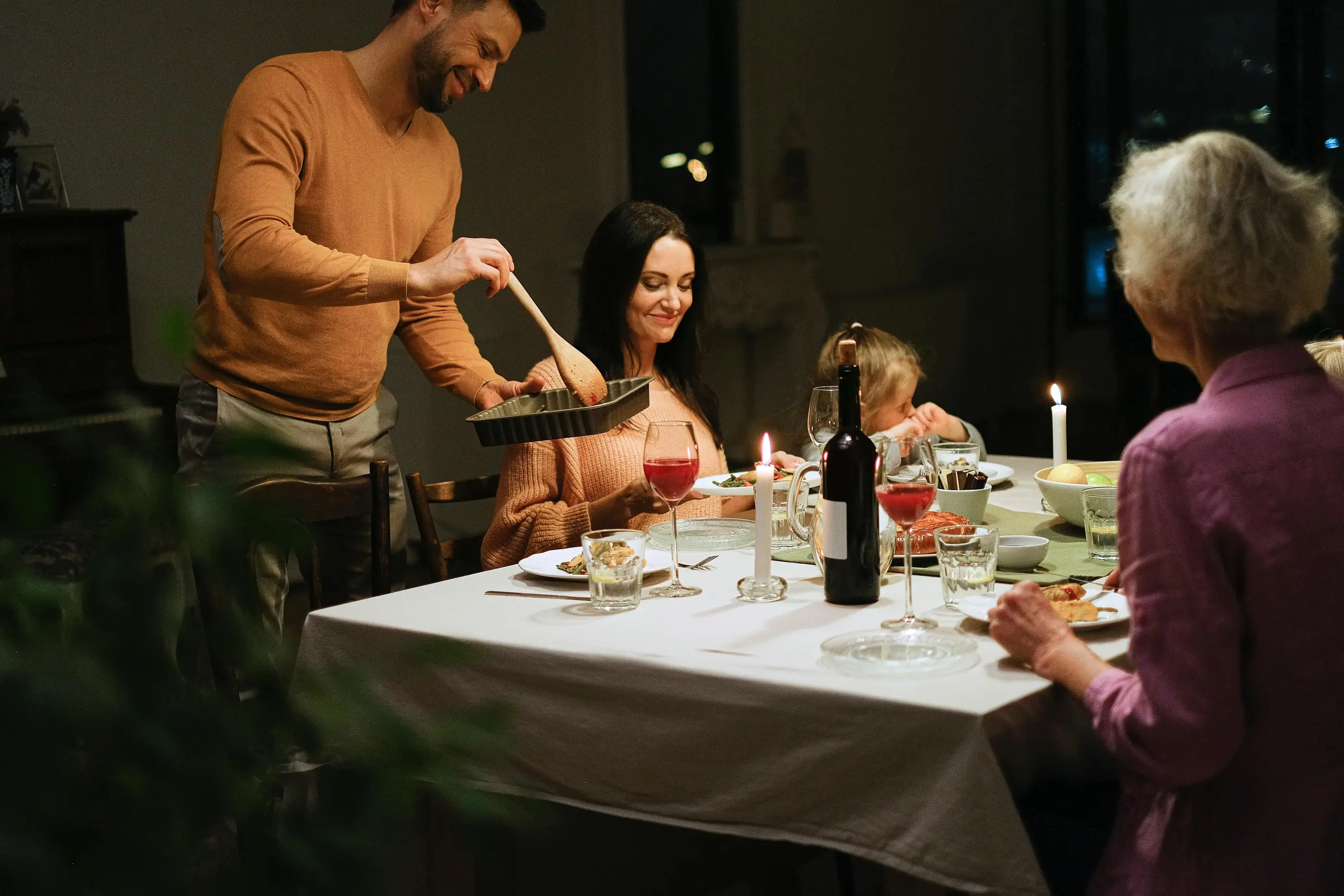 Family gathered around dining table with wine and candles sharing a meal in a cozy setting.