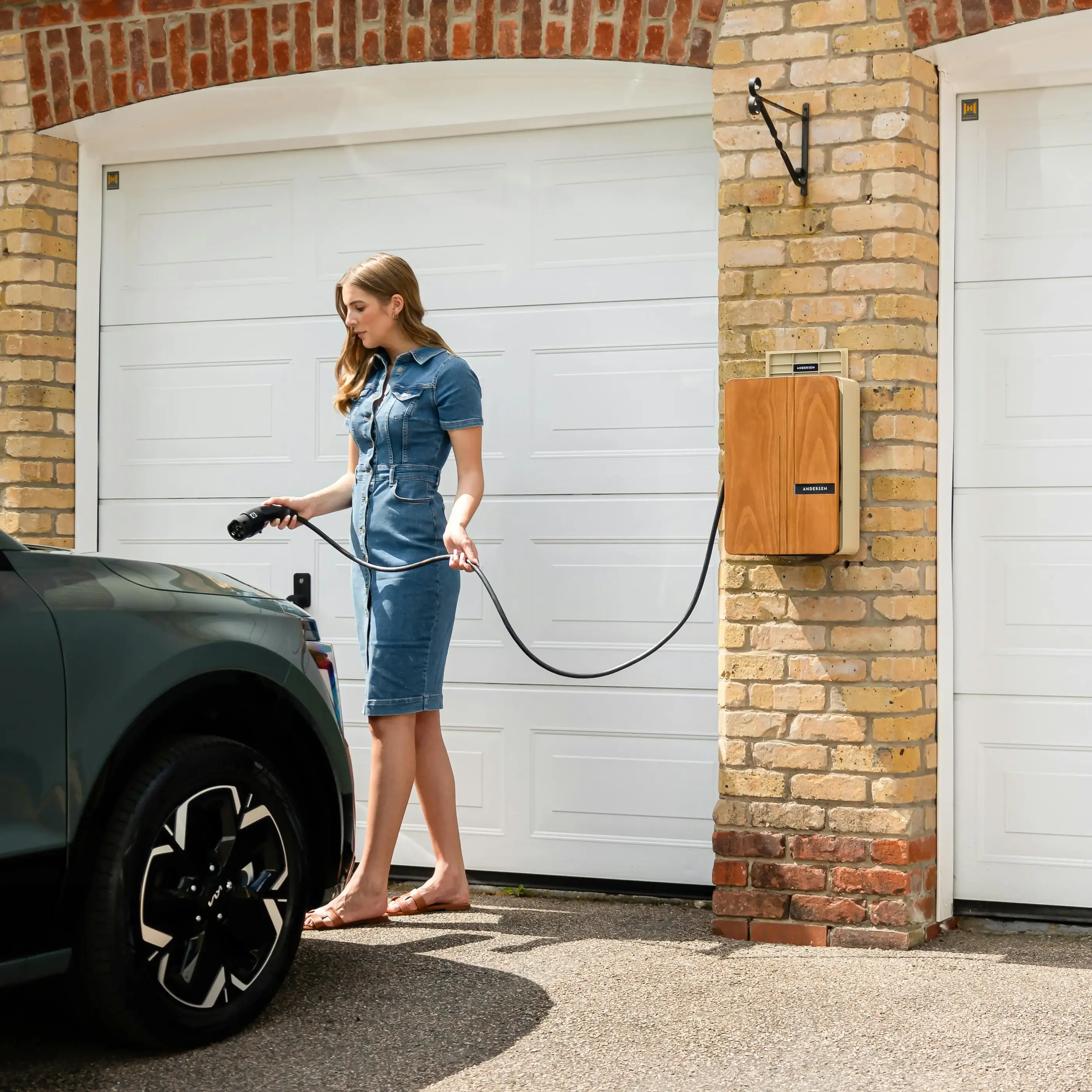 Woman in denim dress holding an electric car charger near a parked vehicle by a garage.