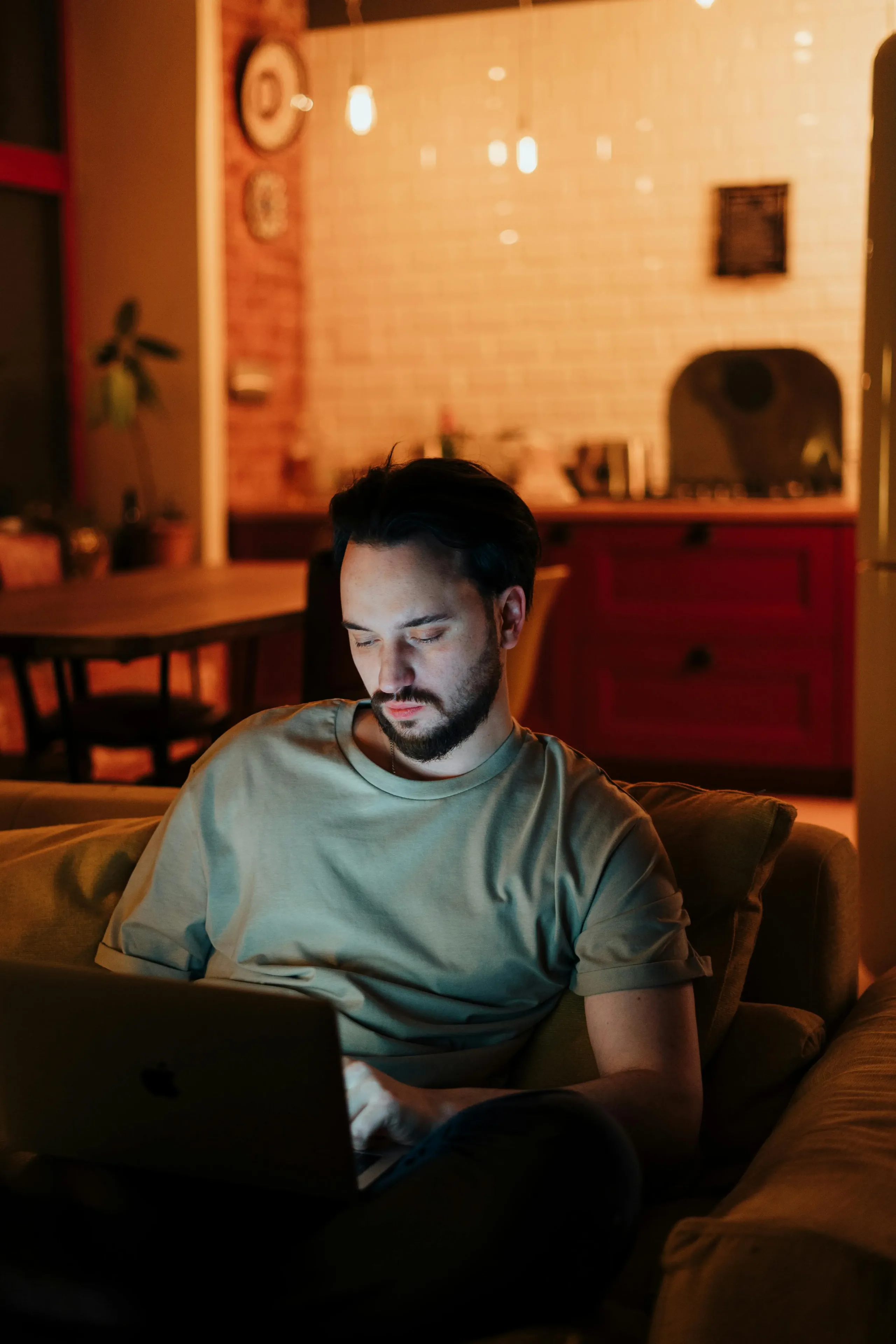 Man with beard sitting on a couch working on a laptop in a warmly lit living room at night.
