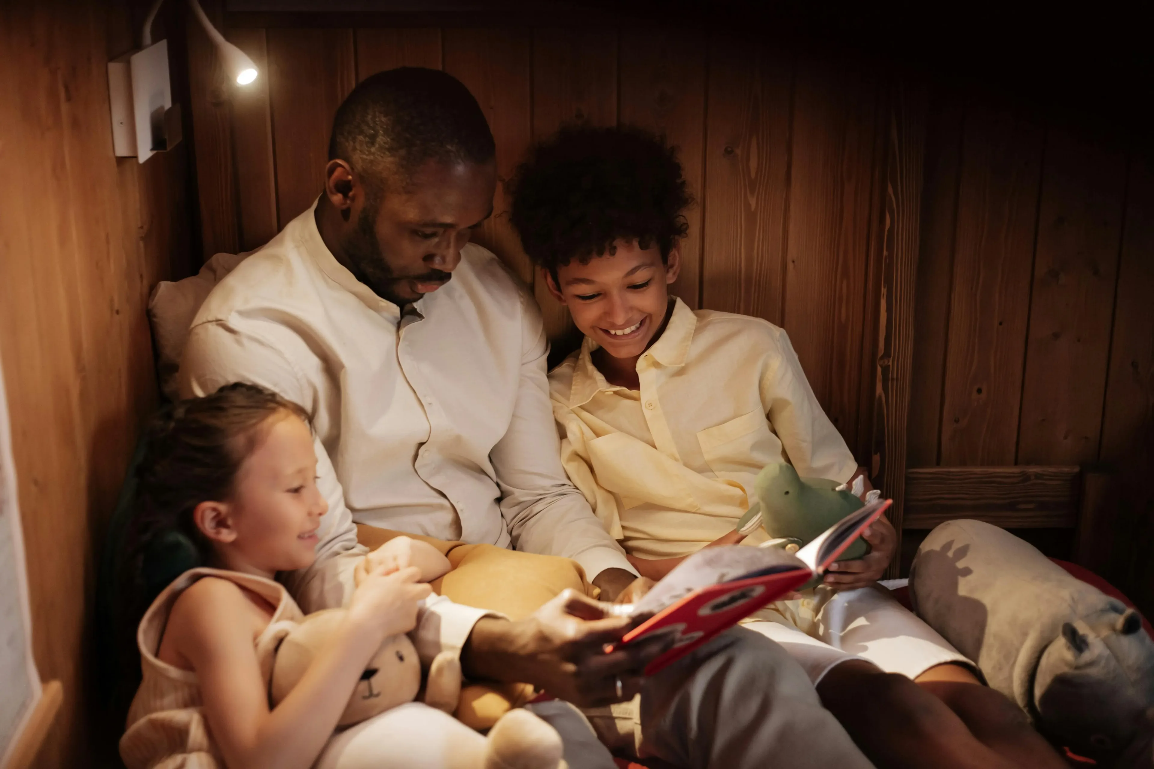 Father reading a book to two smiling children holding stuffed animals in a cozy wooden room.
