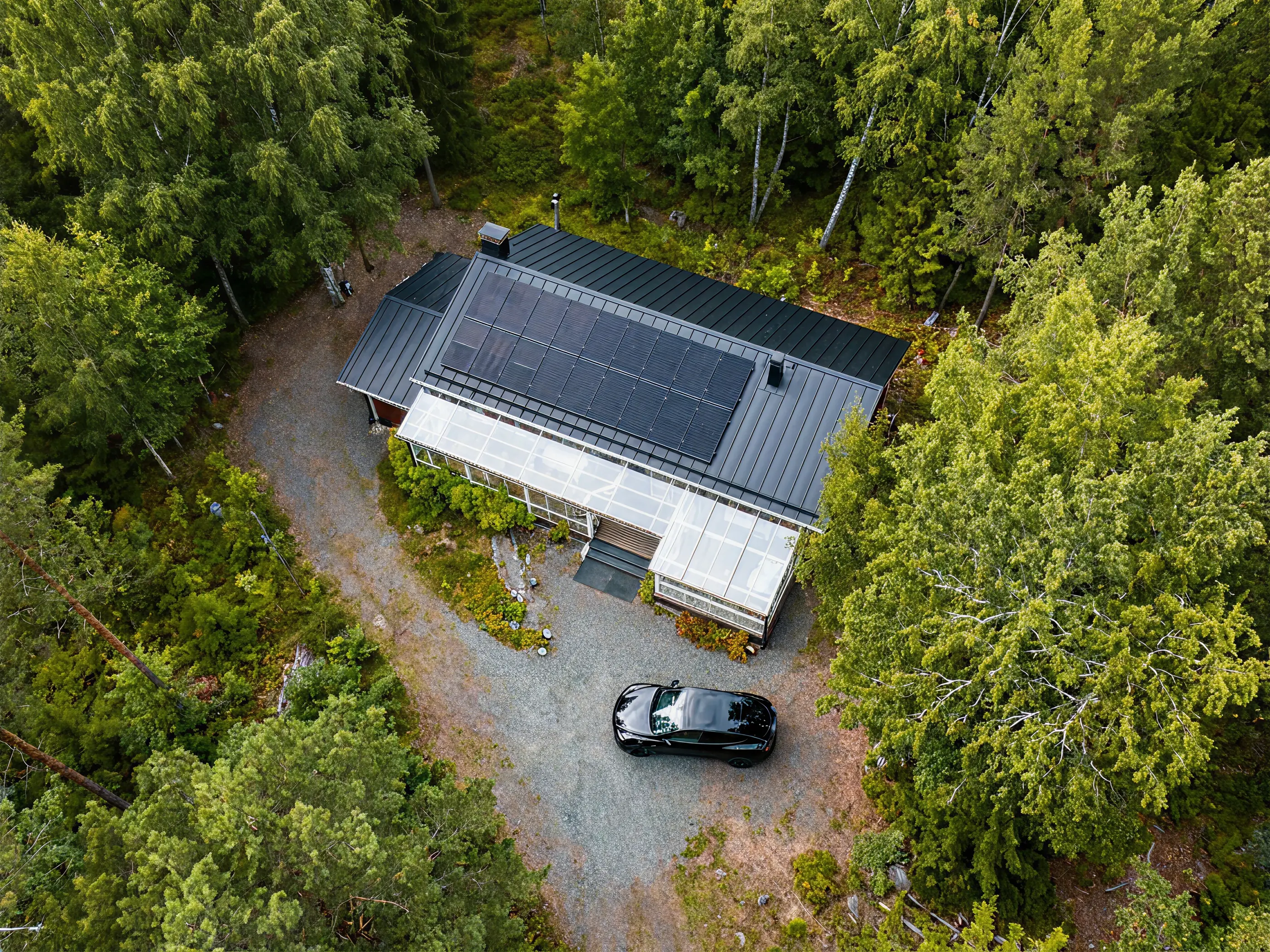 A black car parked on gravel beside a modern house with solar panels, surrounded by dense green trees.