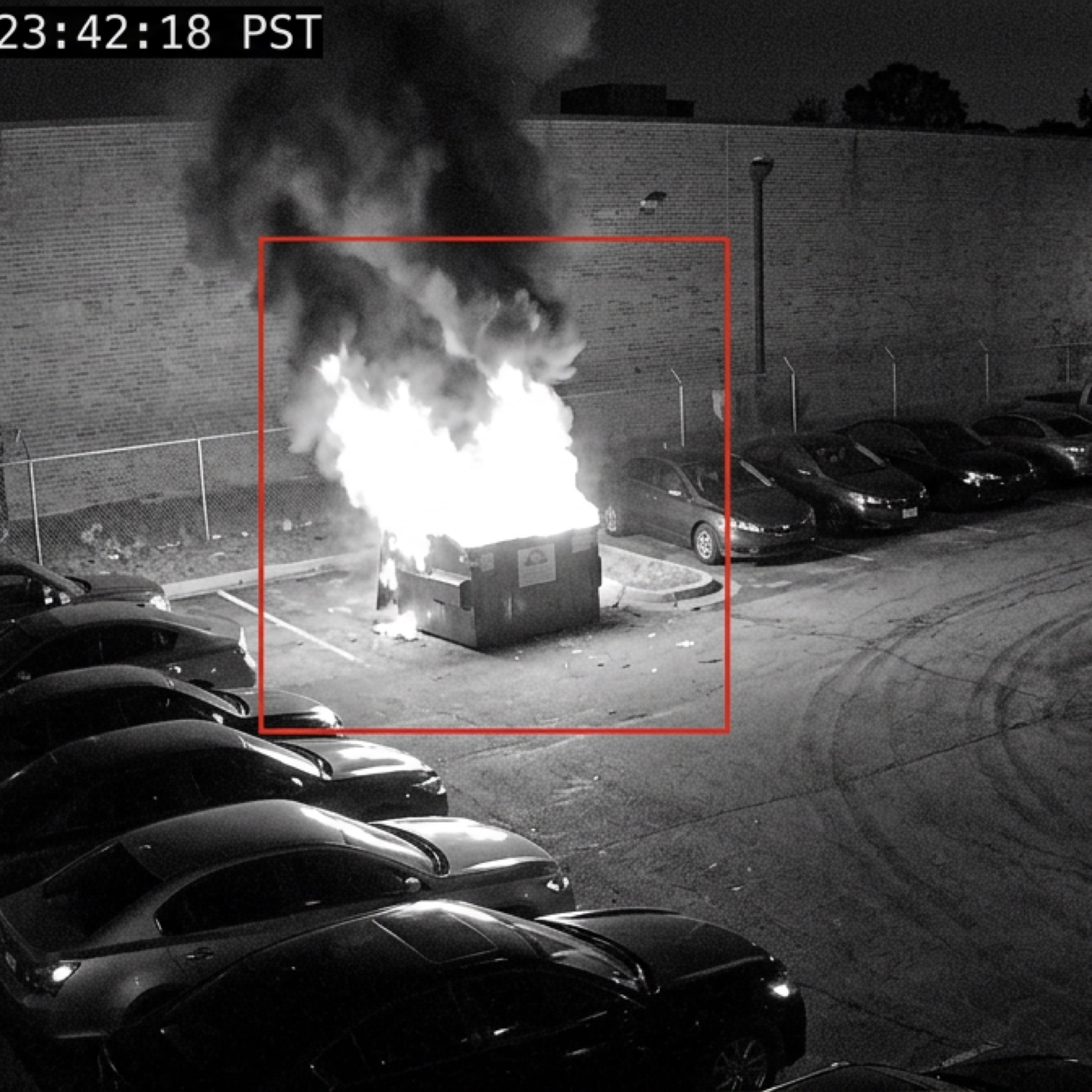 Monochrome night camera footage of a car park, a skip fire burning brightly and ringed by a red detection box.