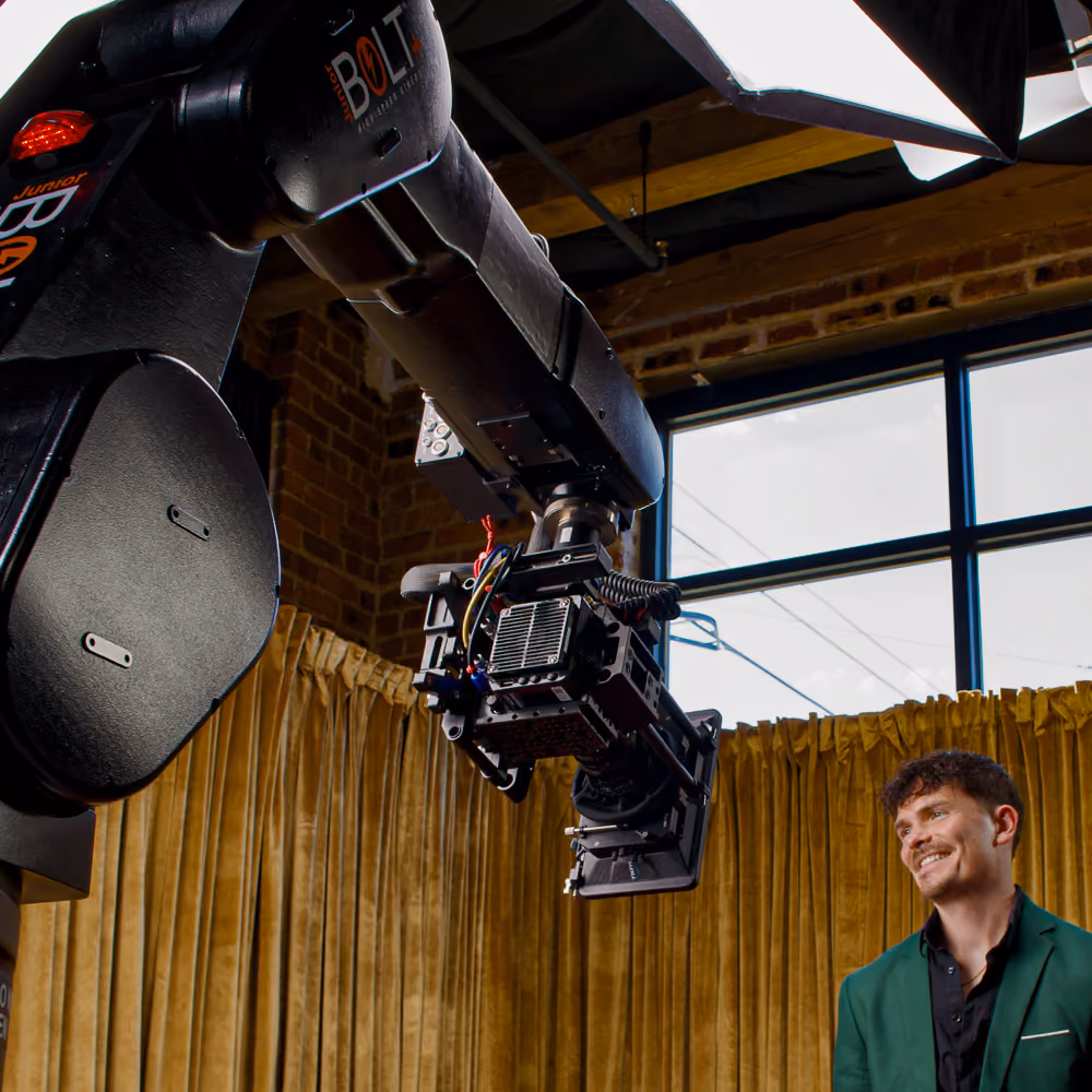 A man in a dark green blazer and black shirt smiling indoors near a glambot robotic arm equipped with a camera, against a backdrop of gold curtains and a window with a brick wall.
