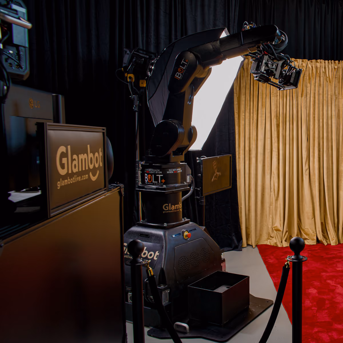 Glambot robotic arm labeled BOLT holding a slowmotion camera, positioned on a platform behind black stanchions with a red carpet and gold curtain backdrop, in a dimly lit studio setup with Glambot branding.