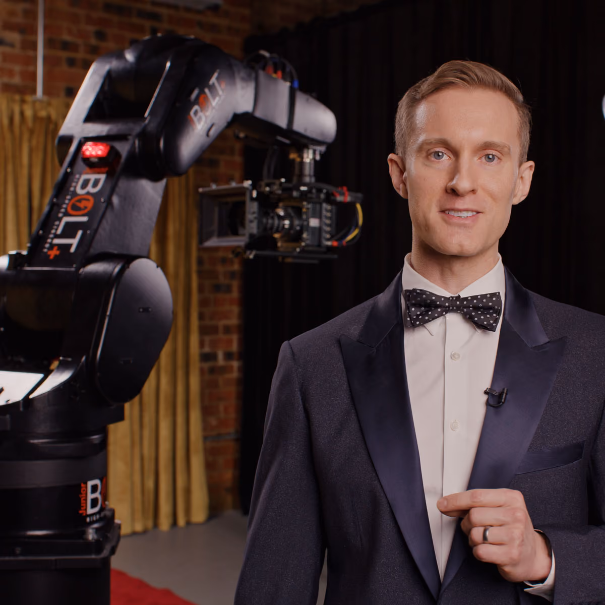 A man in a dark tuxedo with a white shirt and black polka-dot bow tie stands in front of a glambot robotic camera.