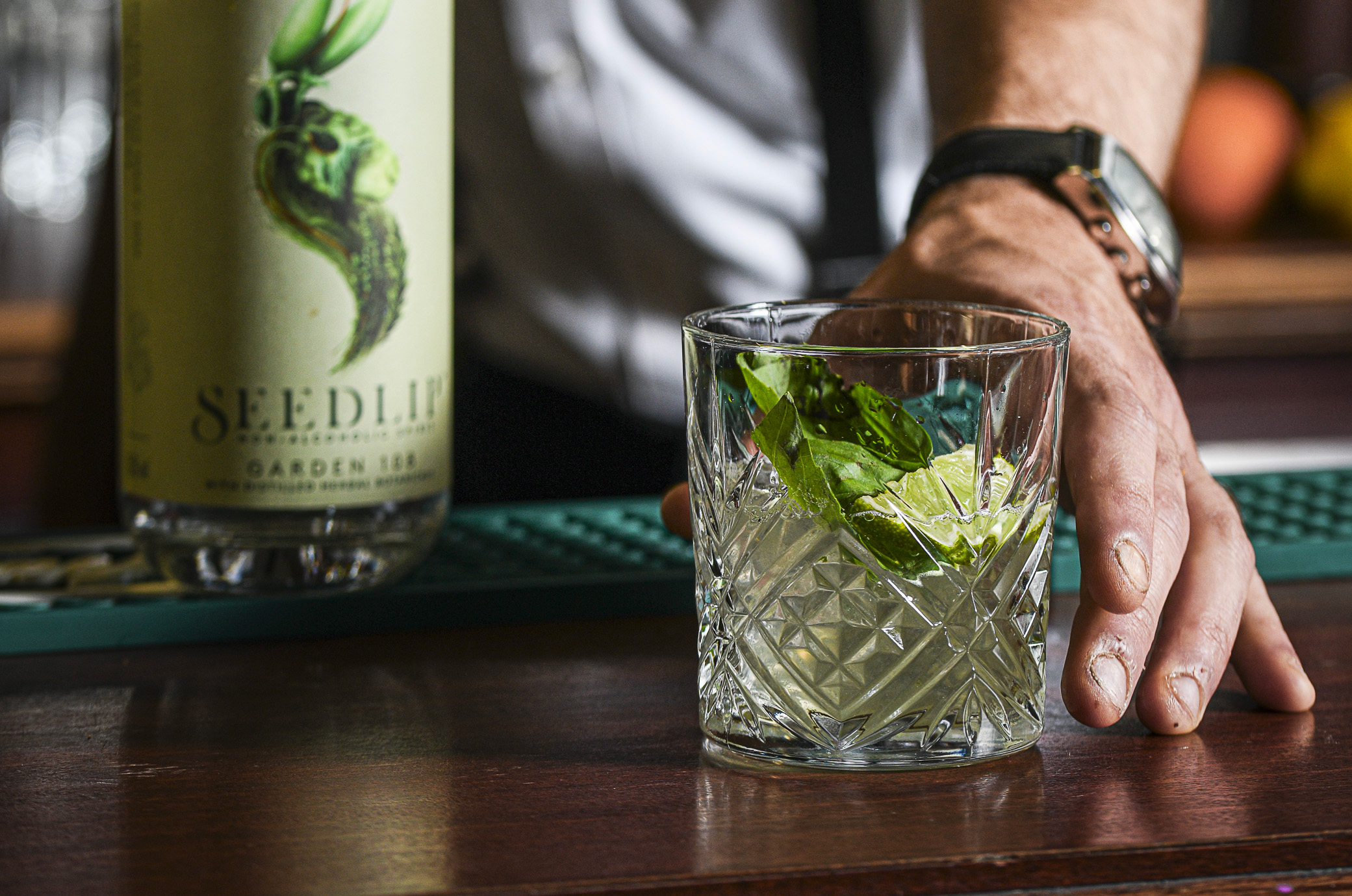 A close-up of a hand resting on a wooden bar counter near a cut-crystal glass containing a clear drink garnished with fresh green leaves and lime wedges, with a bottle labeled Seedlip Garden 108 partially visible in the background.