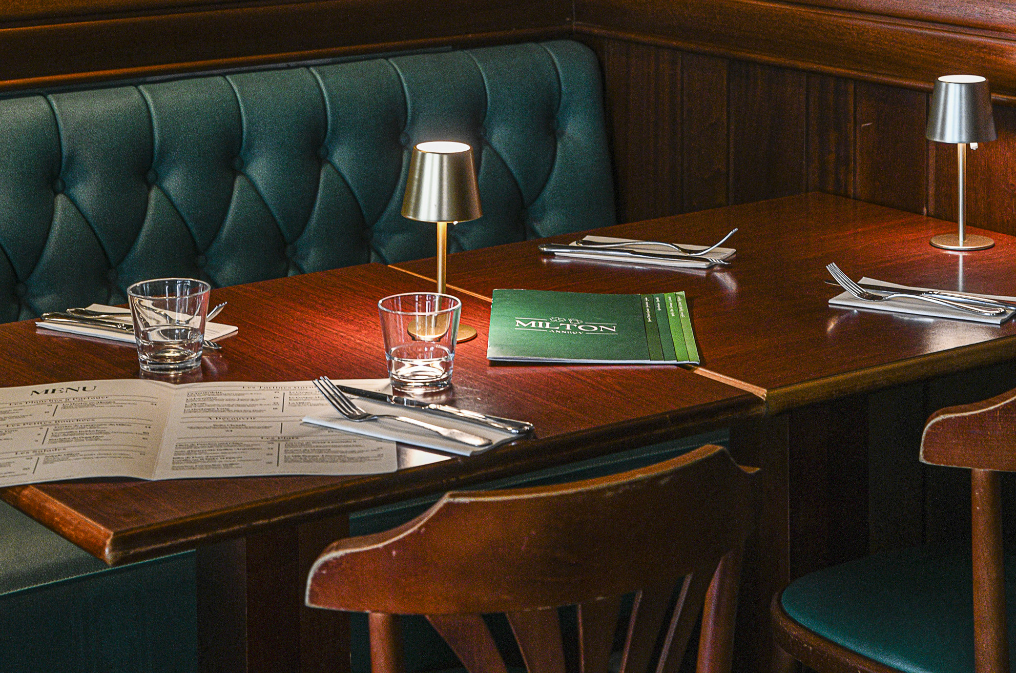 Restaurant table set with water glasses, folded napkins with cutlery, menus, two small modern table lamps, green tufted bench seating, and wooden chairs against wooden paneled walls.