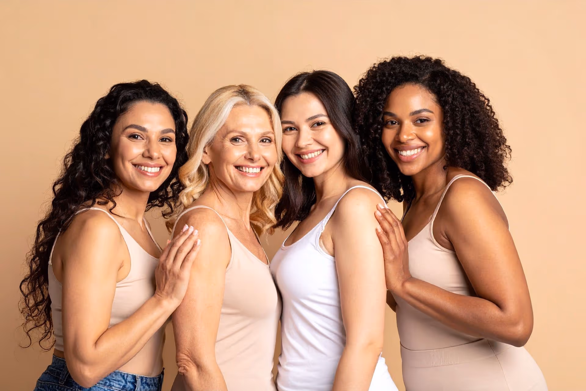 Four diverse women of different ages smiling and standing close together against a beige background.