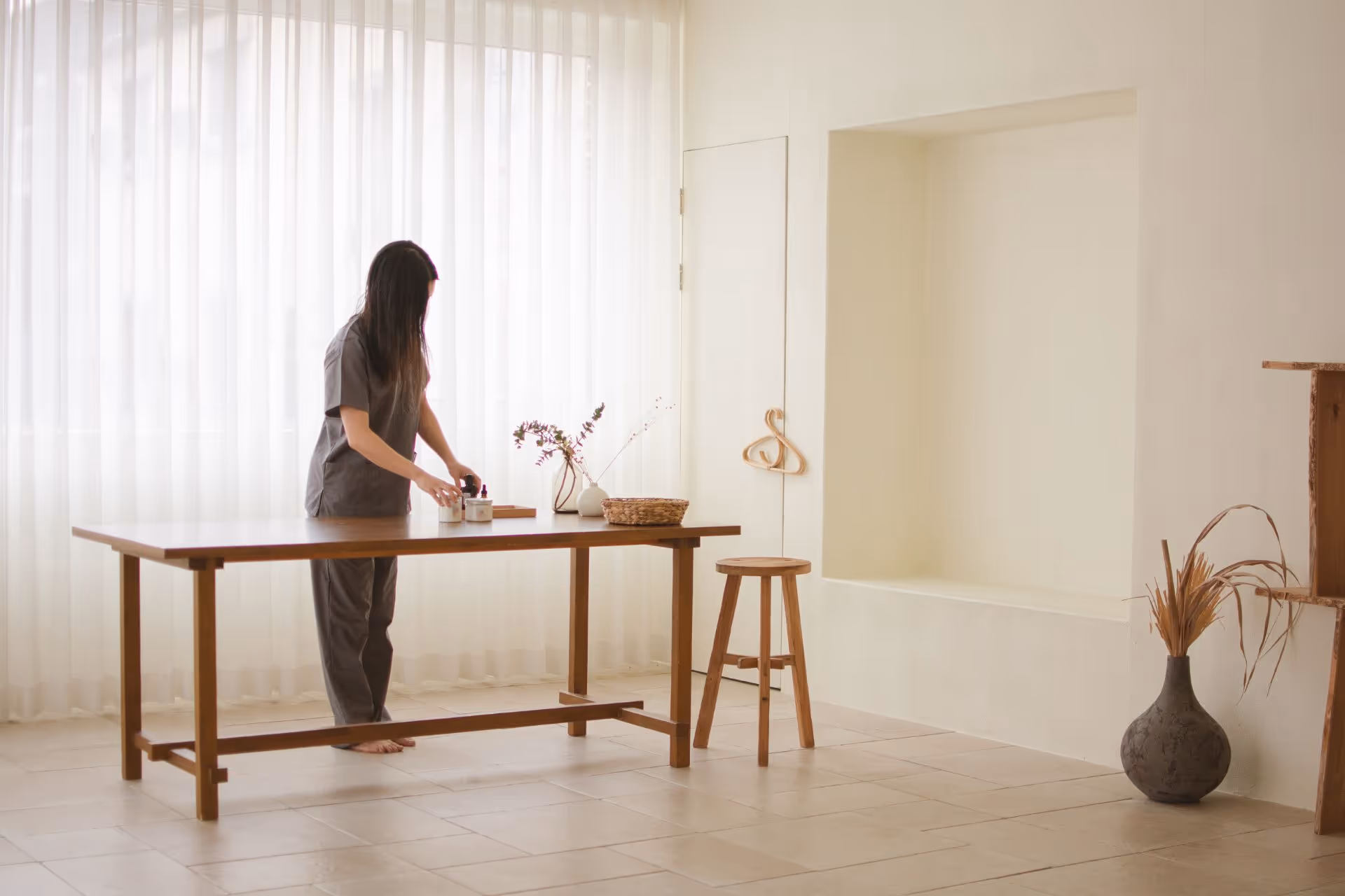 Woman arranging jars and bottles on a wooden table in a minimalist, sunlit room with sheer curtains and natural decor.