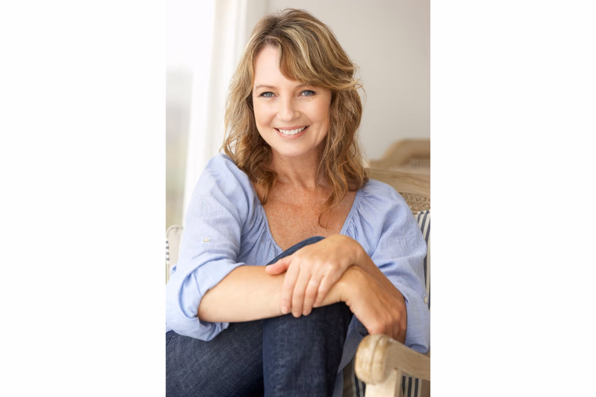 Smiling middle-aged woman with wavy blond hair sitting with arms resting on her knee.