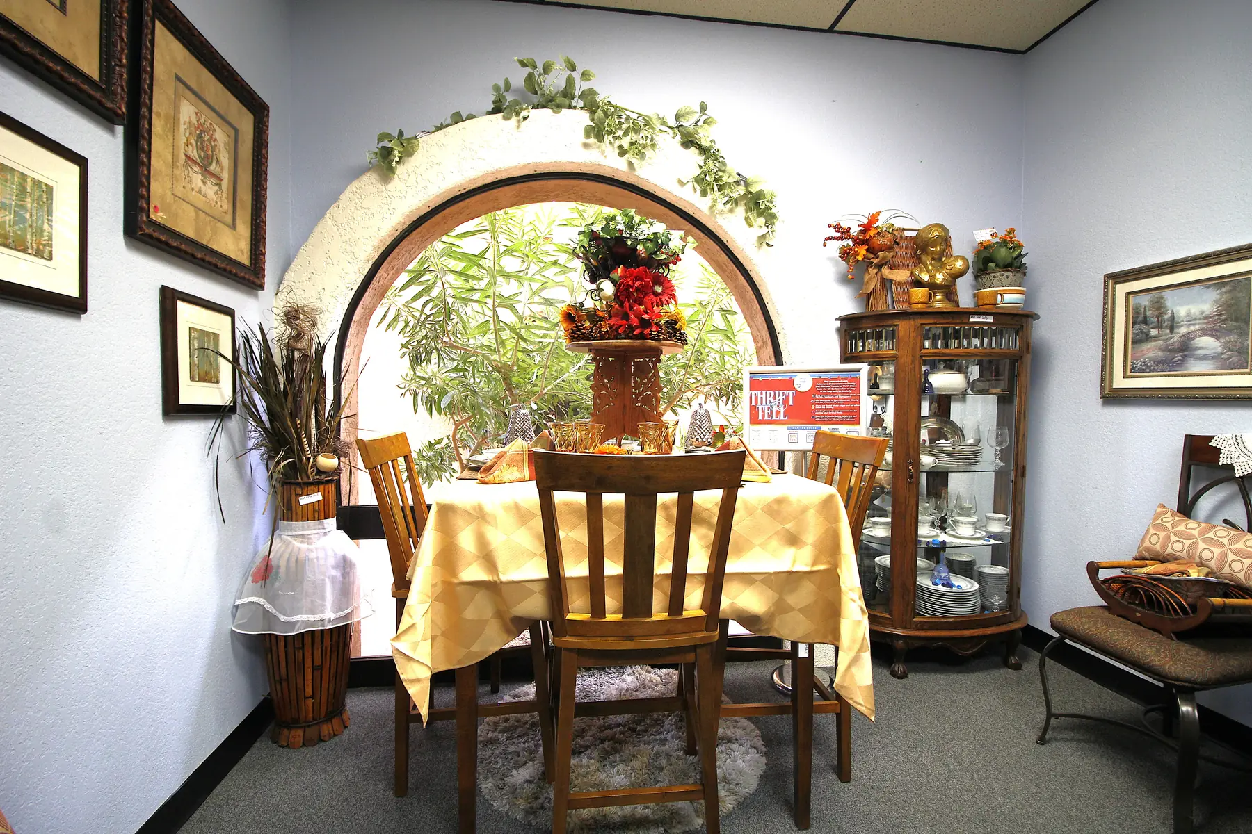 A sunlit dining vignette at Glorious Treasures Thrift Store: a set table beneath an arched window framed with greenery, beside a lit curio cabinet