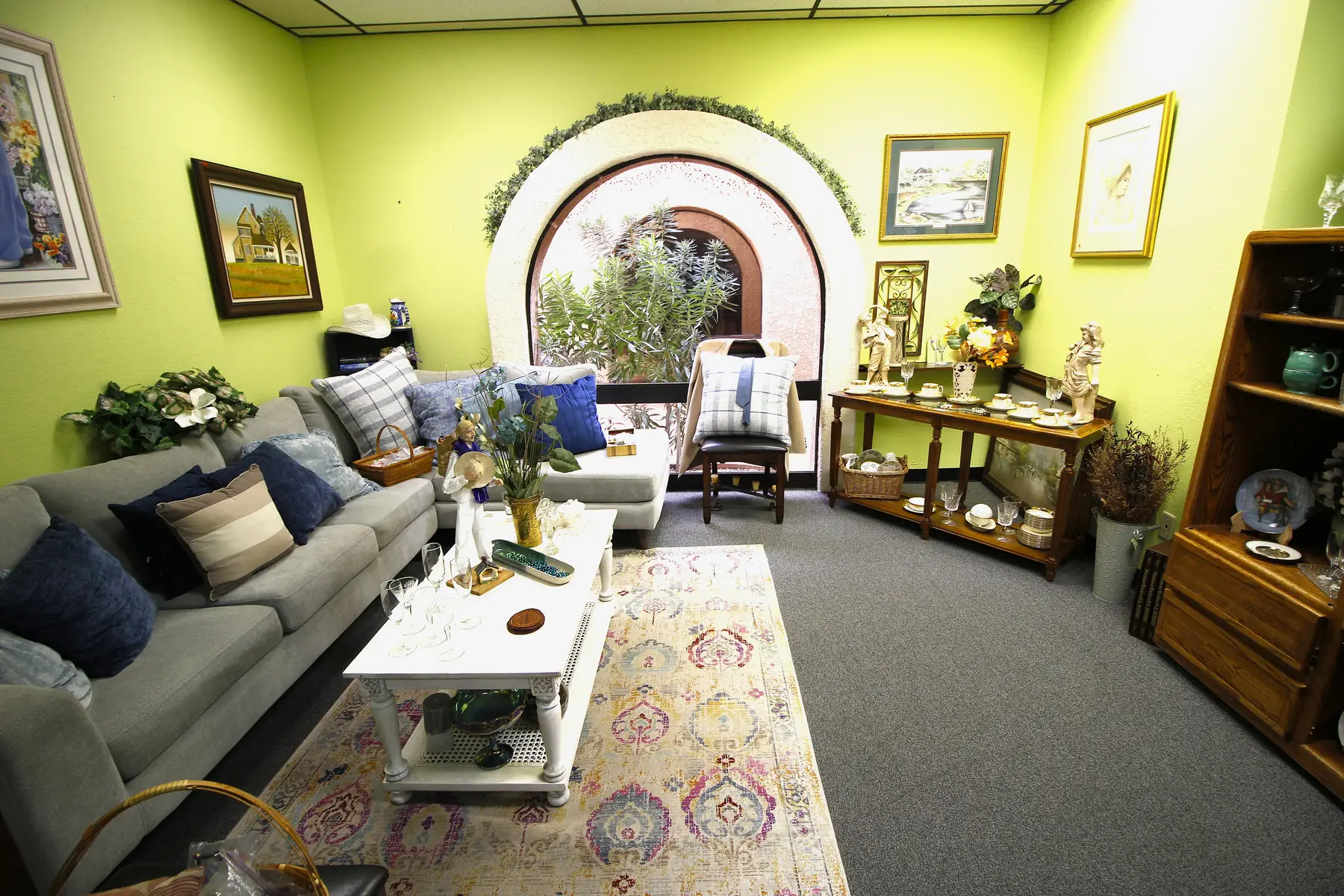 A styled living-room vignette at Glorious Treasures with a gray sectional, white coffee table and patterned rug beneath an arched window