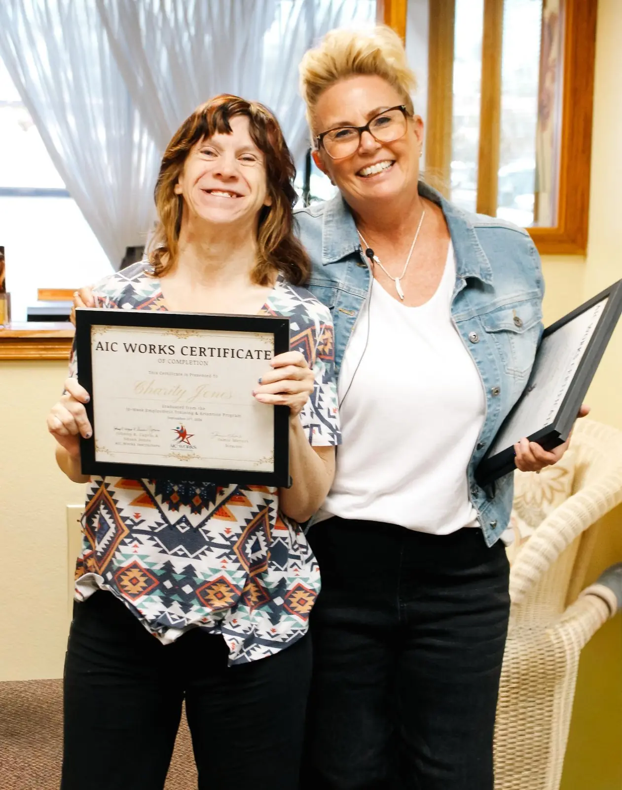 An AIC WORKS graduate holds up her framed Certificate of Completion beside a smiling AIC staff member