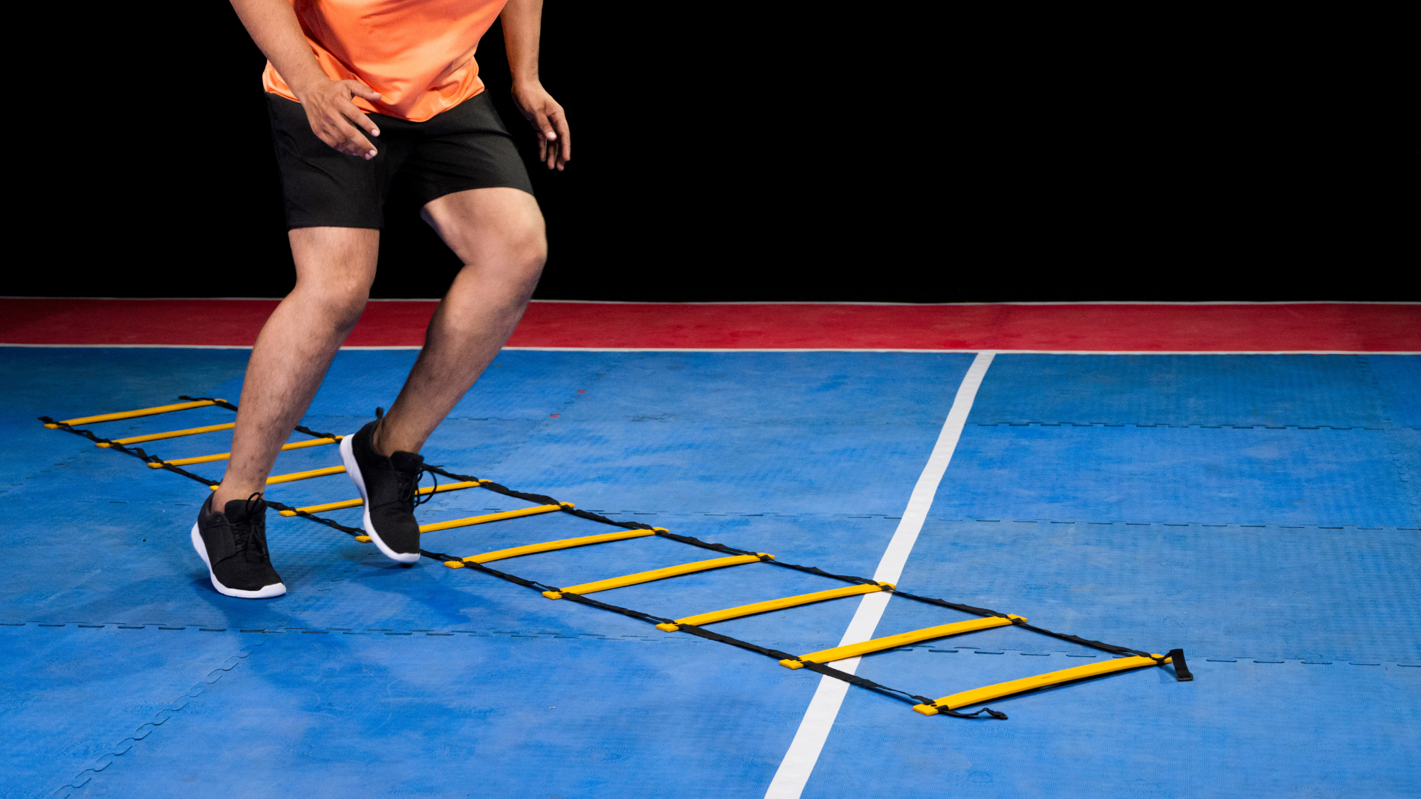 Person wearing black shorts and black athletic shoes with white soles performing agility ladder drills on a blue padded floor with a red border and black background.