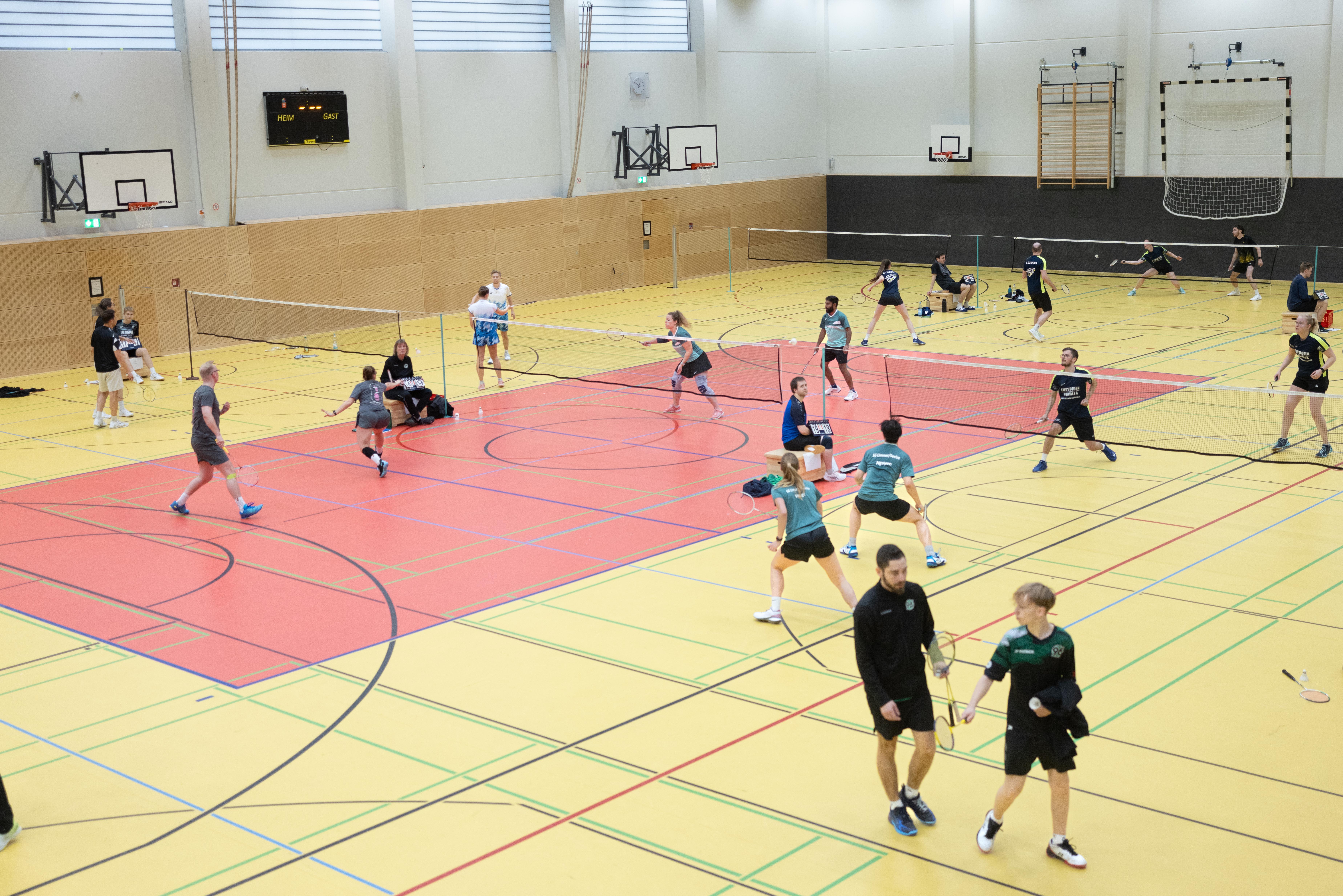 Indoor sports hall with multiple badminton courts where several players are actively playing and practicing badminton, some sitting and watching, with badminton nets and rackets visible.